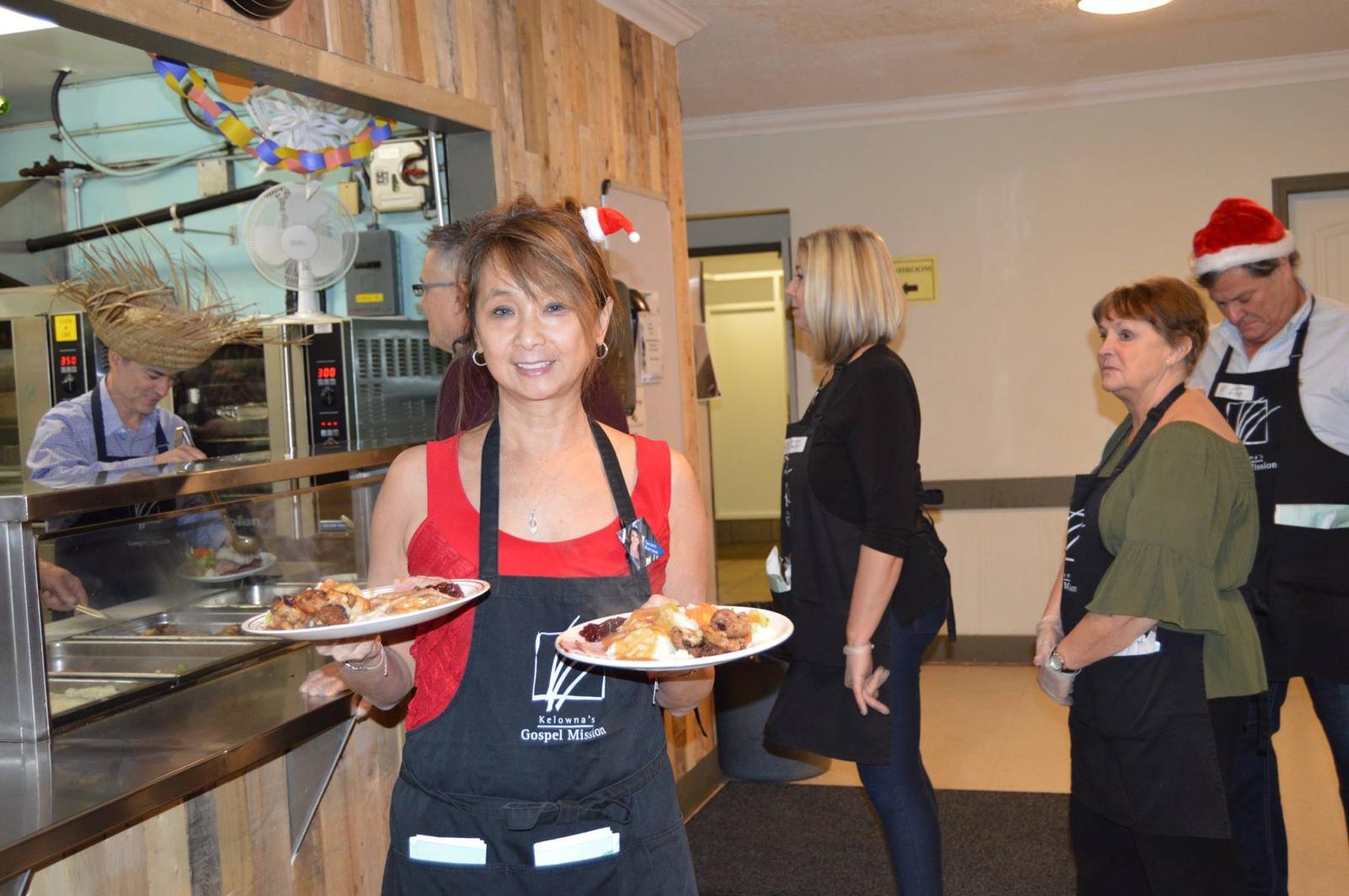 Teresa May carrying two plates of Christmas dinner in a volunteer apron at Kelowna's Gospel Mission in 2019.
